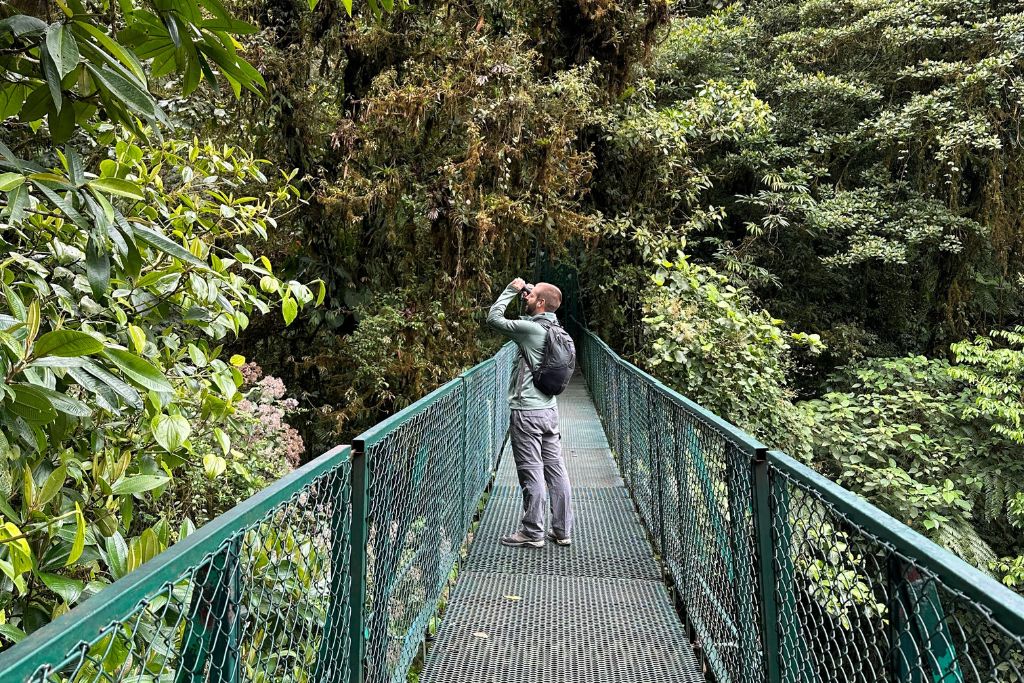 A man on a green hanging bridge in Monteverde cloud forest, looking through a telescope for wildlife