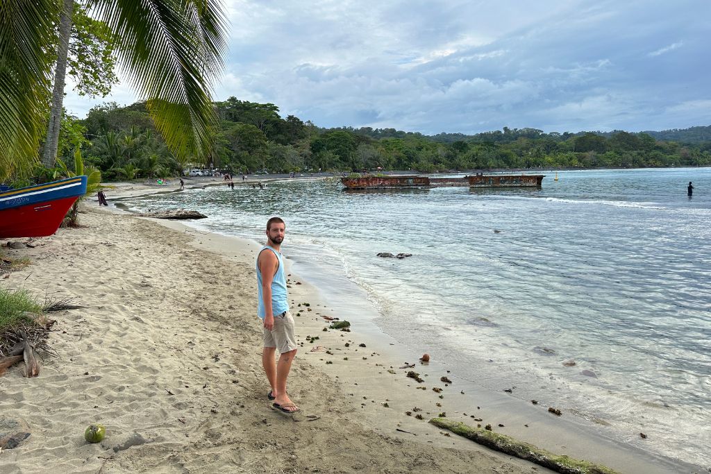 A man standing on a sandy beavh in Puerto Viejo with a ship wreck in the sea behind him