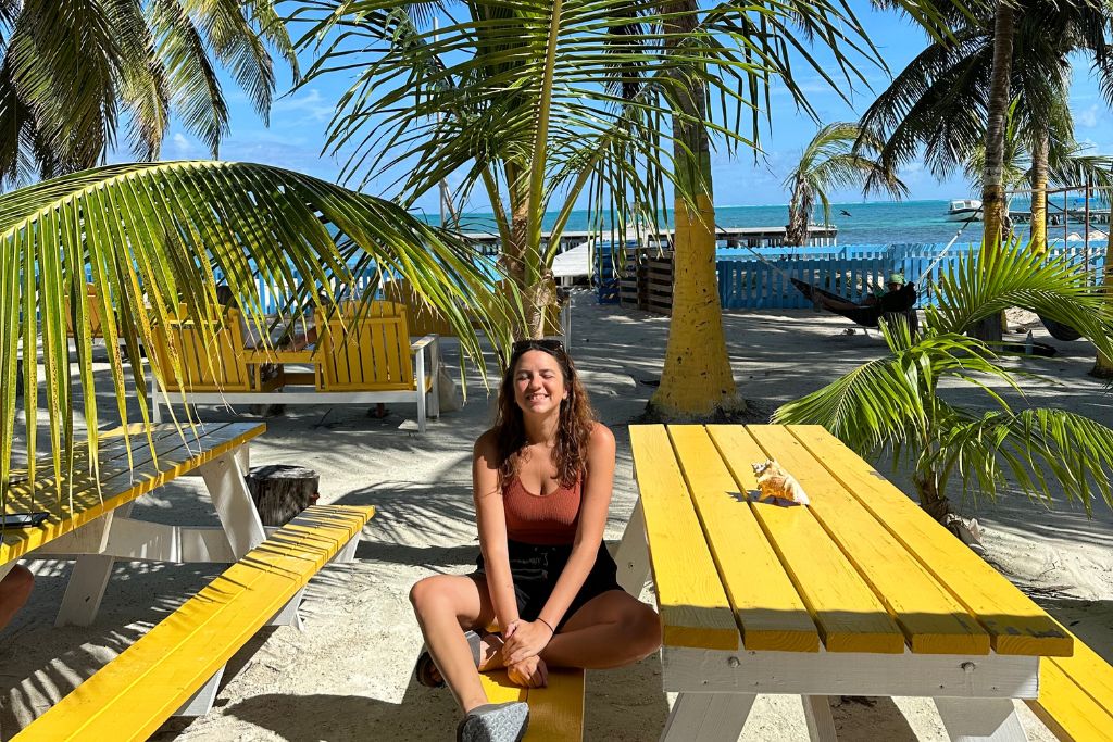 a girl sitting on a yellow picnic bench with palm trees behind her in Caye Caulker